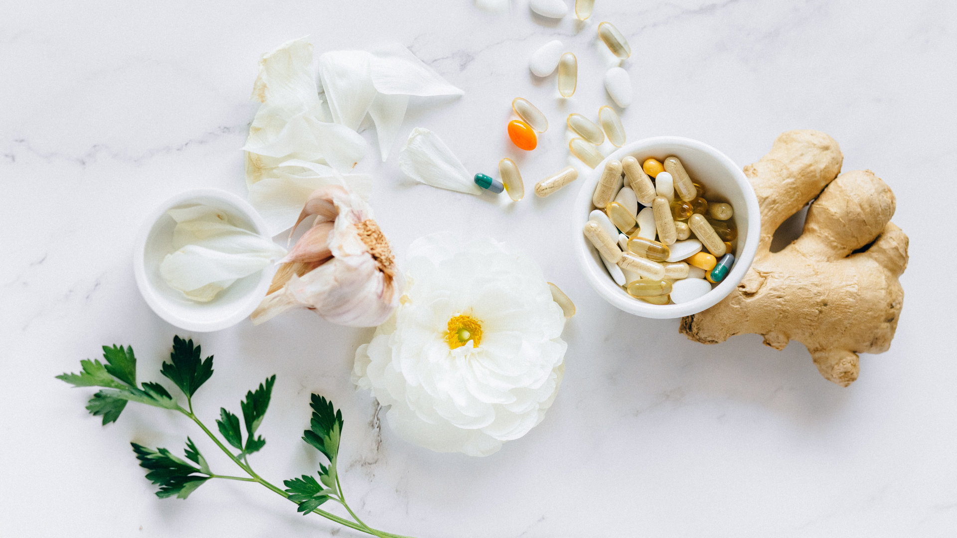 Welby Wellness & Aesthetics flat lay with a white ranunculus flower, fresh ginger, and an assortment of capsules and tablets in white bowls on a marble surface, accented by parsley and a soft, clean aesthetic