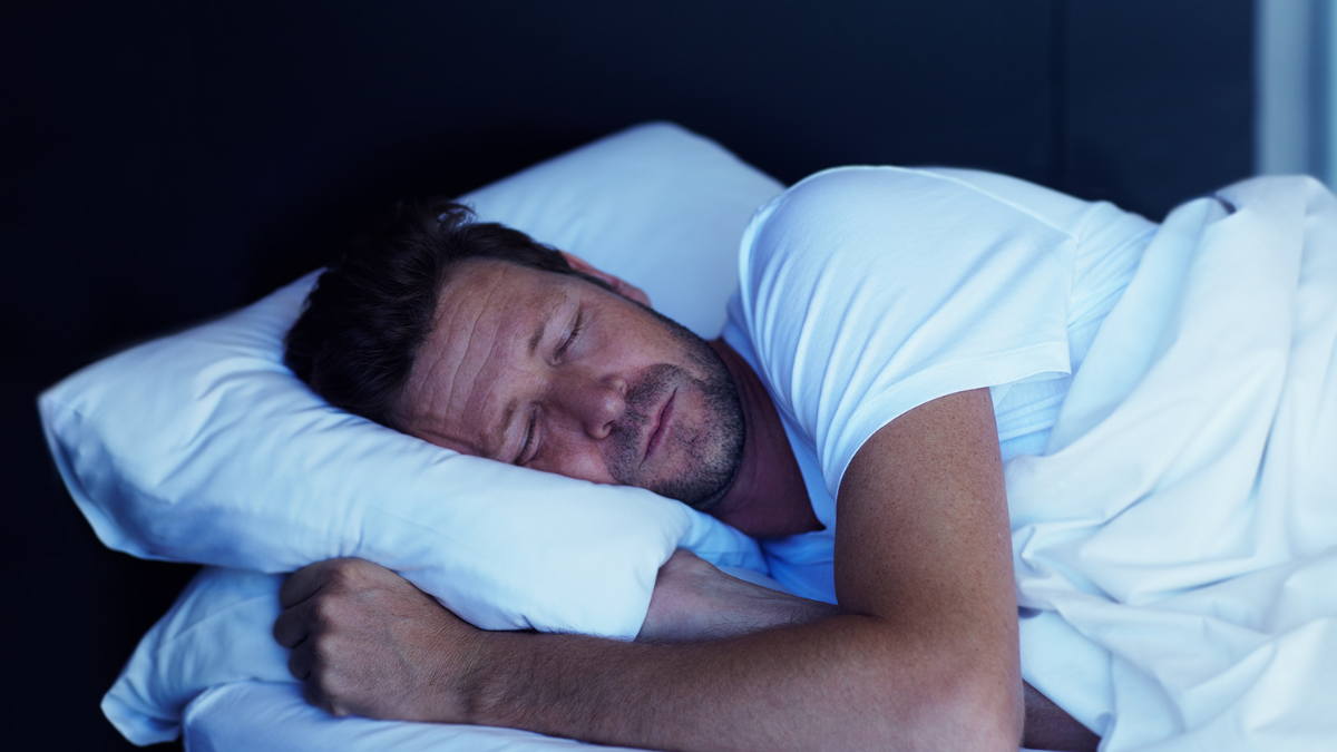 Man with short hair sleeping on a white pillow in a dark room, wearing a white t-shirt.