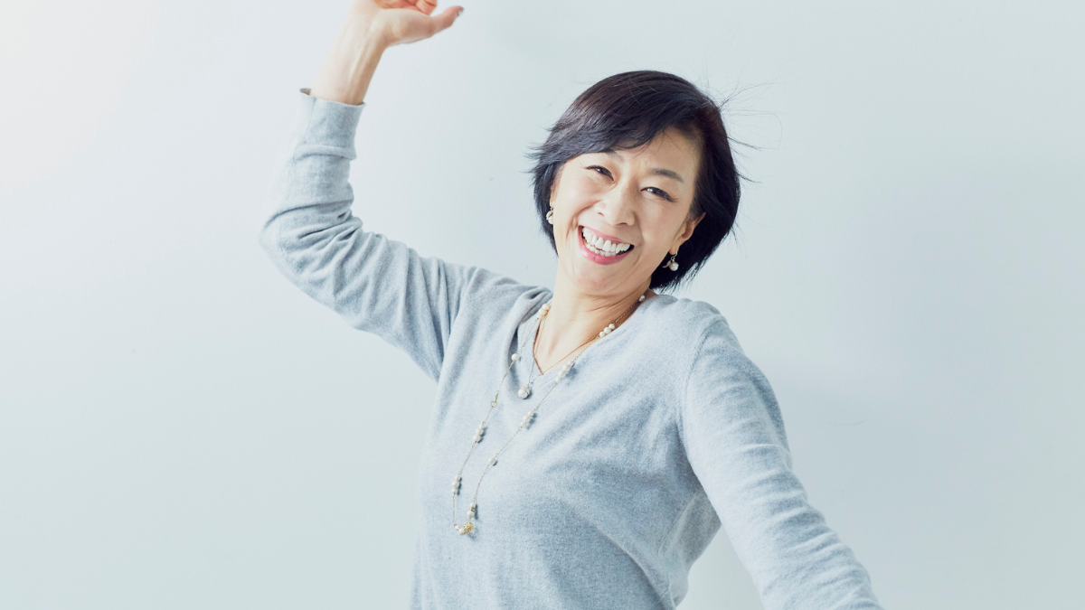 Woman with short black hair wearing a gray sweater, smiling and raising her arm in a joyful pose against a light background.