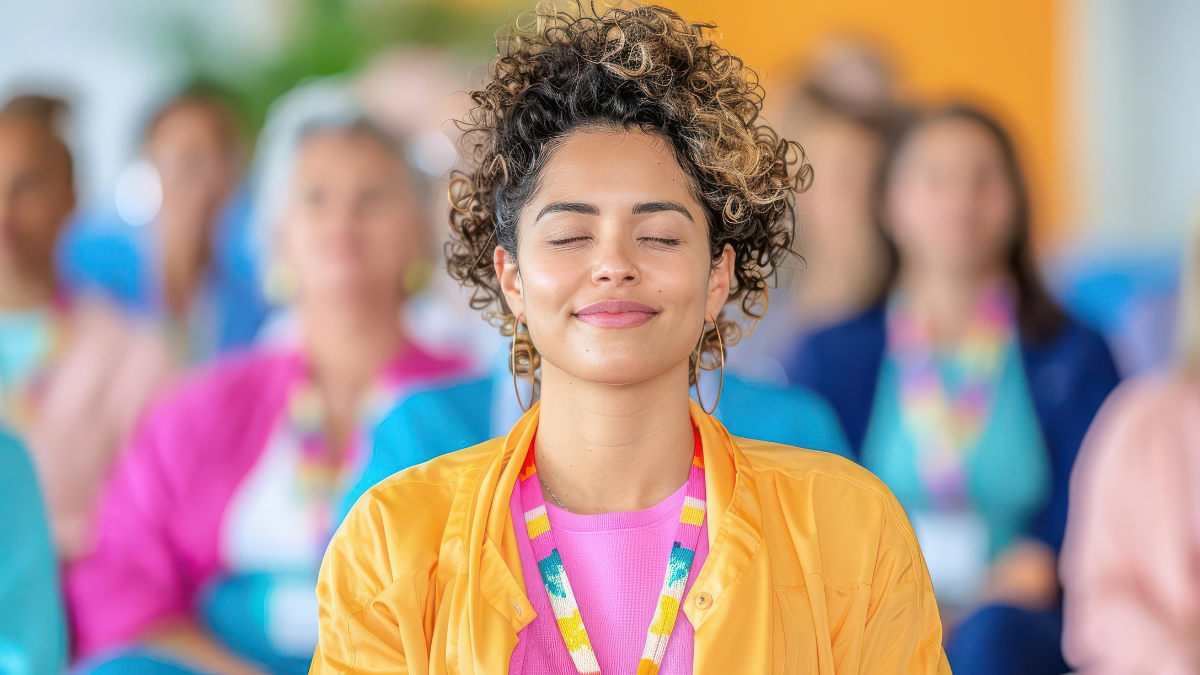 Woman with curly hair wearing a yellow jacket and pink shirt, eyes closed, meditating in a group of people.