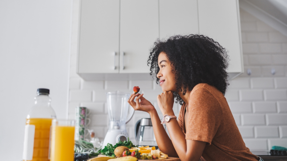 Woman with curly hair enjoying a piece of fruit while sitting at a kitchen table with a blender and drinks.