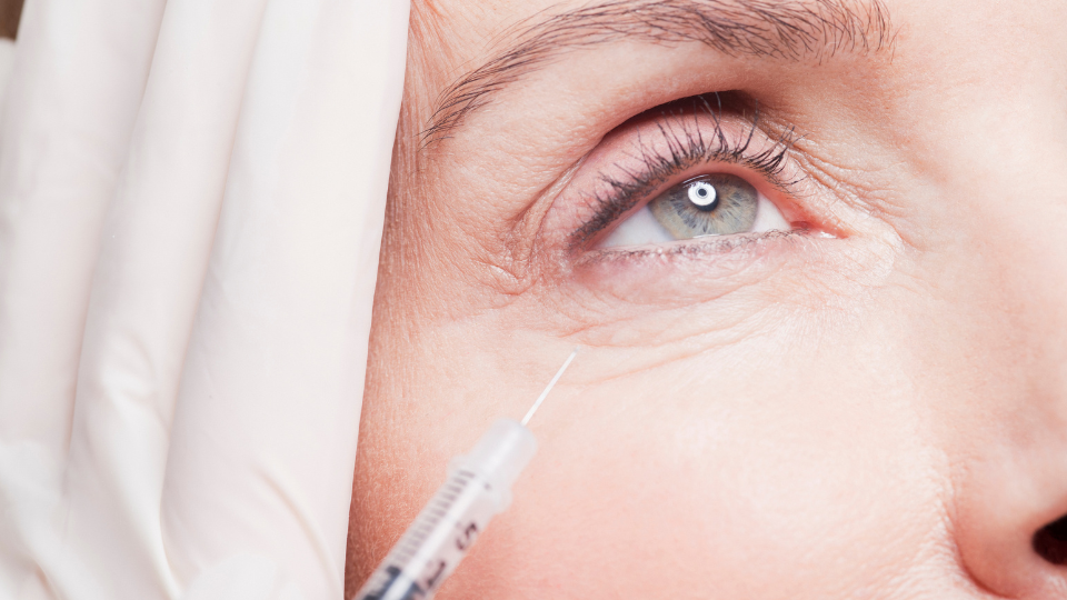 Close-up view of a person's eye with a syringe being held near the under-eye area for cosmetic treatment.