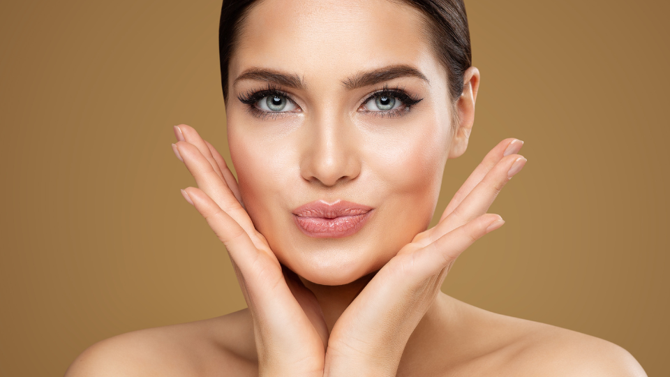 Cheek lift aesthetic: a woman with glowing skin and blue eyes, hands framing her face, against a warm tan background, highlighting dermal filler options like Juvederm and Restysl—Allergan.