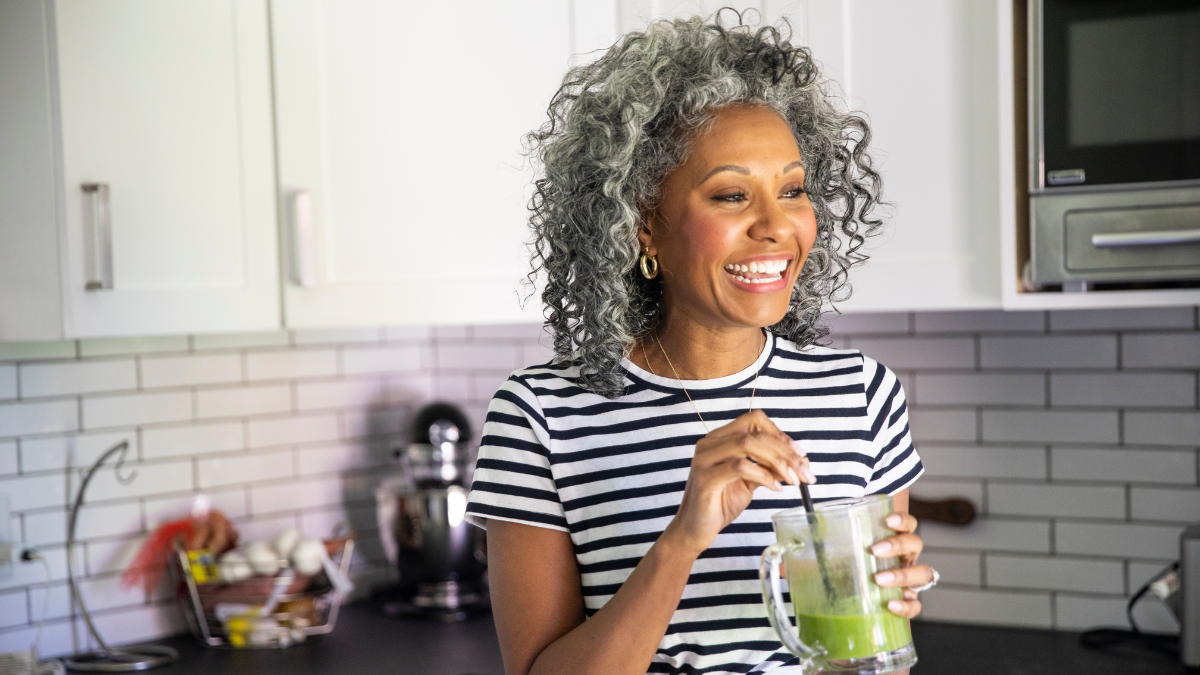 Woman with curly gray hair smiling while holding a blender with a green smoothie in a kitchen.