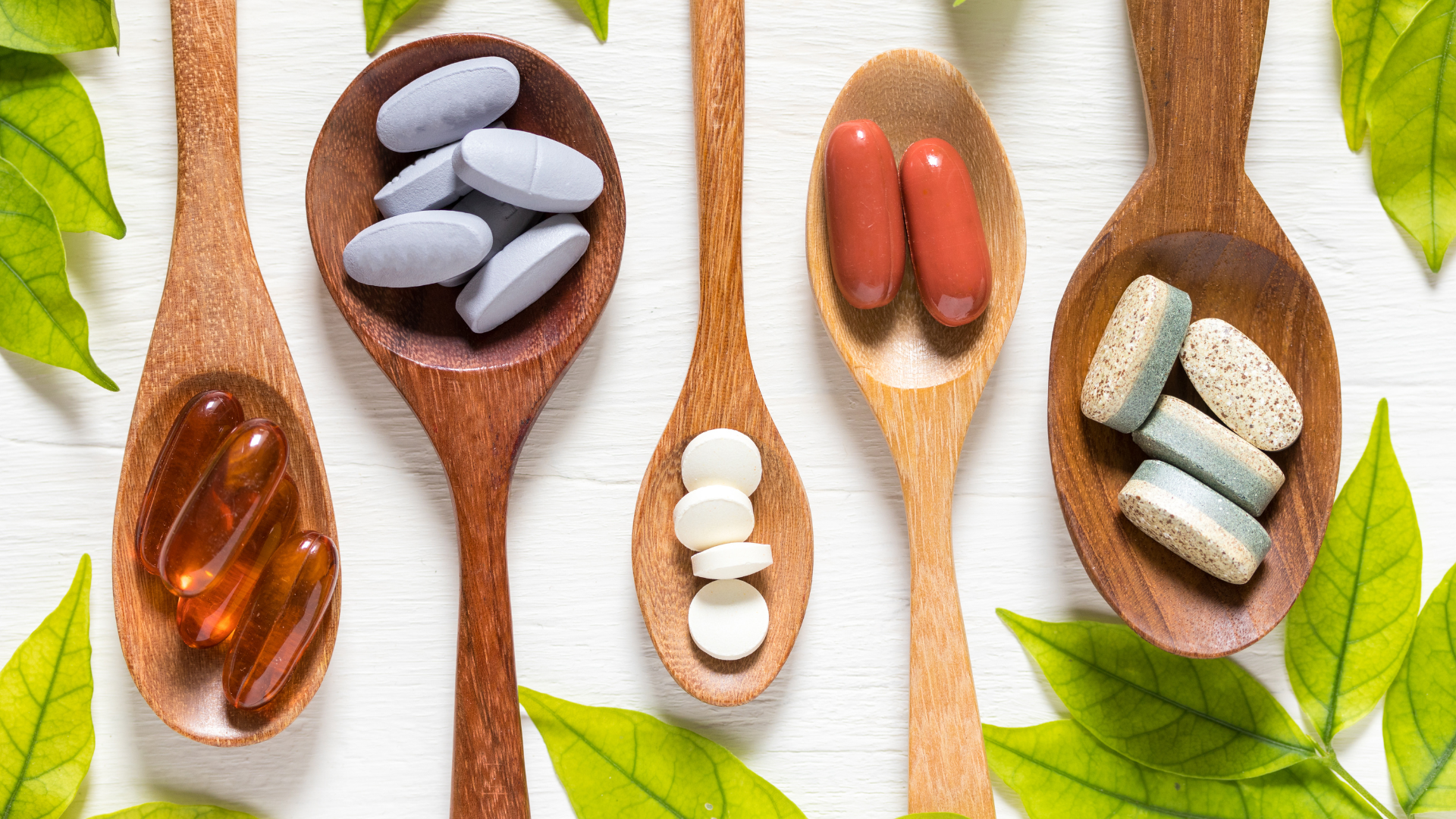 Assorted capsules and tablets in wooden spoons laid out on a white surface, featuring blue-gray, white, red, and beige pills amid fresh green leaves