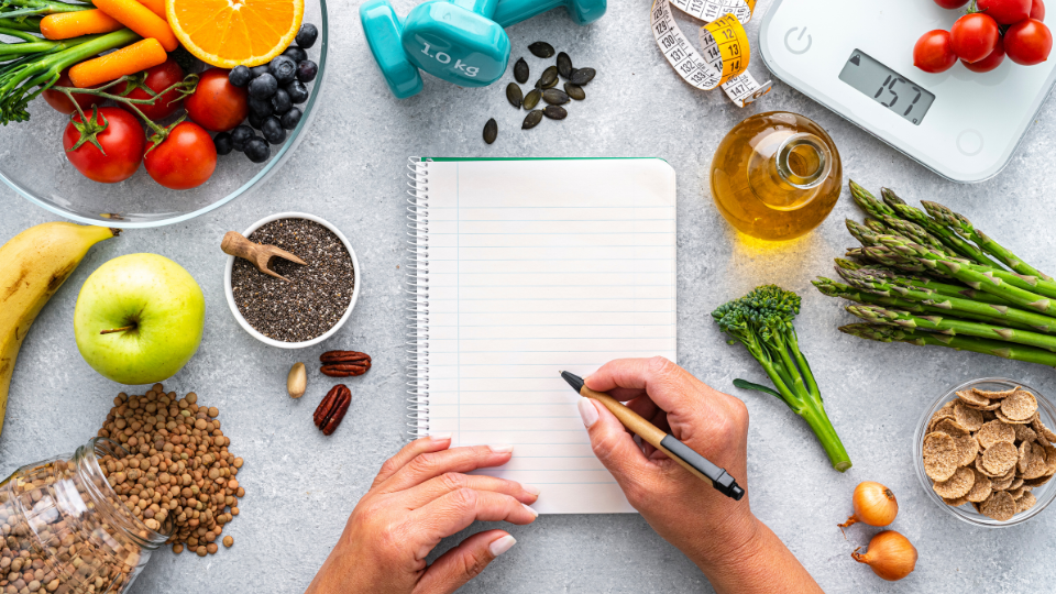 A flat lay of various healthy foods, exercise weights, and a notebook with a pen for planning.